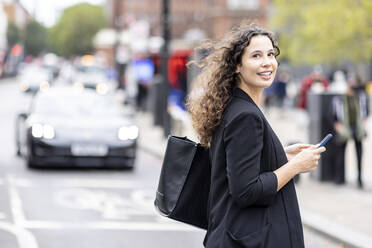 Lächelnde Geschäftsfrau mit Mobiltelefon beim Überqueren der Straße in der Stadt - WPEF05747