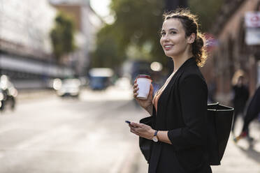 Nachdenkliche Geschäftsfrau mit Smartphone und Einweg-Kaffeebecher auf der Straße - WPEF05766