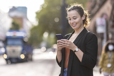 Lächelnde Geschäftsfrau mit Mobiltelefon in der Stadt - WPEF05769