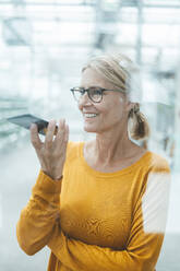 Geschäftsfrau, die eine Sprachnachricht auf ihrem Mobiltelefon sendet, gesehen durch das Glas eines Büros - JOSEF06725