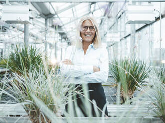 Cheerful scientist standing amidst plants at greenhouse - JOSEF06735