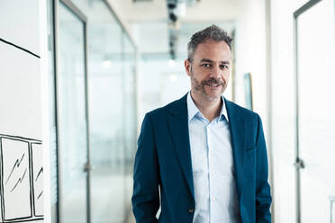 Smiling businessman standing in corridor at office - JOSEF06836