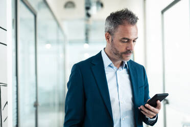 Businessman using smart phone in corridor at office - JOSEF06837