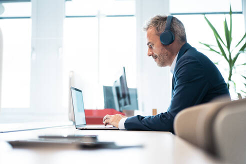 Businessman wearing wireless headphones using laptop at desk in workplace - JOSEF06839