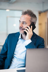 Smiling businessman sitting with laptop at desk talking on smart phone - JOSEF06844