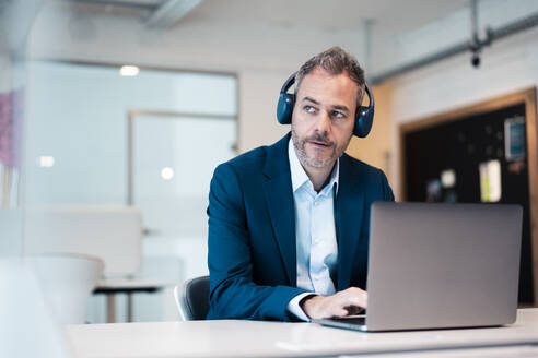 Thoughtful businessman wearing wireless headphones using laptop at desk in workplace - JOSEF06846