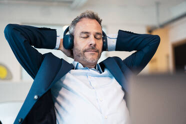Relaxed businessman with hands behind head wearing wireless headphones in office - JOSEF06849