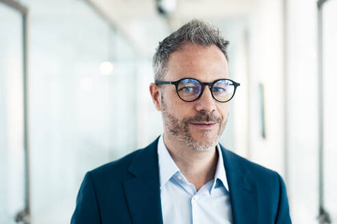 Confident businessman wearing eyeglasses in corridor at office - JOSEF06857