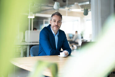 Businessman sitting with coffee cup at desk in workplace - JOSEF06864