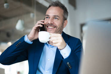 Lächelnder Geschäftsmann, der eine Kaffeetasse hält und mit einem Mobiltelefon spricht - JOSEF06866