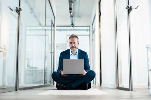 Selbstbewusster Geschäftsmann im Schneidersitz mit Laptop im Bürokorridor - JOSEF06871