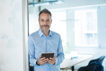 Businessman leaning on doorway with tablet computer at office - JOSEF06872