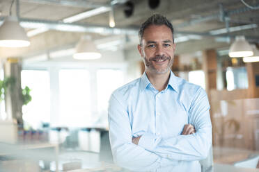 Smiling businessman standing with arms crossed at workplace - JOSEF06873