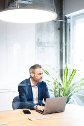 Confident businessman sitting with laptop at desk in workplace - JOSEF06877