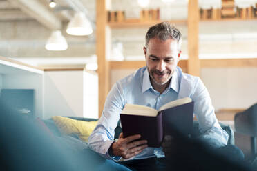 Smiling businessman reading book sitting at workplace - JOSEF06878