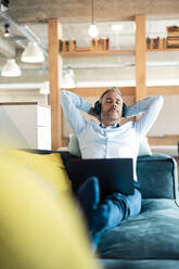 Relaxed businessman listening music sitting with laptop on sofa at office - JOSEF06880