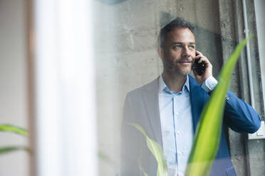 Thoughtful businessman talking on mobile phone seen through window - JOSEF06882