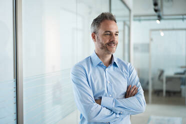 Smiling businessman with arms crossed standing in corridor at office - JOSEF06894