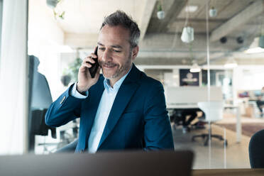 Smiling businessman talking on mobile phone looking at laptop in workplace - JOSEF06895