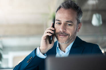 Geschäftsmann, der am Arbeitsplatz mit seinem Handy telefoniert und auf seinen Laptop schaut - JOSEF06896