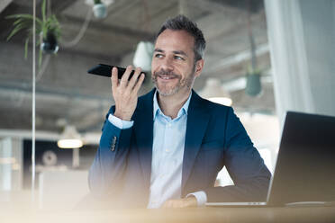 Lächelnder Geschäftsmann, der mit seinem Laptop am Arbeitsplatz sitzt und über sein Handy spricht - JOSEF06898