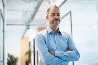 Thoughtful businessman with arms crossed standing in corridor at workplace - JOSEF06900