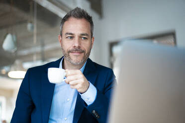 Geschäftsmann hält Kaffeetasse und schaut auf Laptop im Büro - JOSEF06904