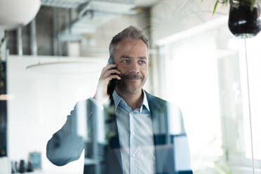 Smiling businessman looking away talking on mobile phone seen through glass wall - JOSEF06911