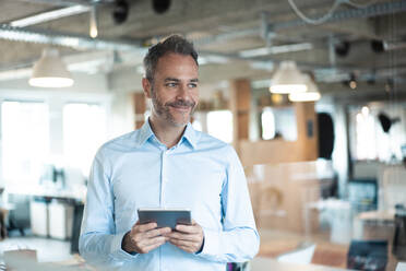 Smiling businessman standing with tablet computer at workplace - JOSEF06916