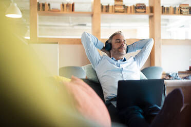 Relaxed businessman listening music sitting with laptop on sofa at office - JOSEF06920
