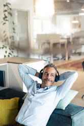 Relaxed businessman listening music sitting on sofa at office - JOSEF06921