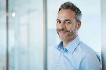 Smiling businessman with stubbles leaning on glass wall in office corridor - JOSEF06928