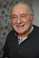 Positiv faltiger älterer Mann mit grauem Haar in Freizeitkleidung, der in die Kamera schaut, während er in einem hellen Raum zu Hause sitzt - ADSF33559