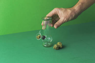 Crop anonymous male hand demonstrating small glass bong placed on green surface with dry cannabis leaves for smoking - ADSF33568