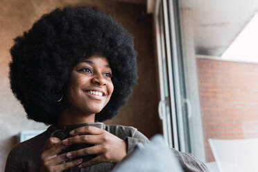 Positive African American female with Afro hairstyle looking away with smile while sitting near window with mug of hot drink - ADSF33577