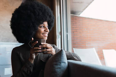 Positive African American female with Afro hairstyle looking away with smile while sitting near window with mug of hot drink - ADSF33578