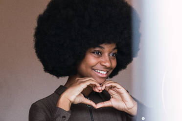 Cheerful African American female blogger with Afro hairstyle making heart gesture with hands and looking at camera through ring lamp - ADSF33579