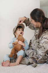 Caring female soldier in uniform putting cap on cute barefoot son with toy while playing in light room at home - ADSF33588