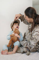 Caring weiblichen Soldaten in Uniform Putting Kappe auf niedlichen barfuß Sohn mit Spielzeug beim Spielen in hellen Raum zu Hause - ADSF33588