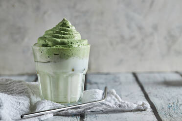 Ein Glas frischer Dalgona-Kaffee mit Matcha steht neben einem Strohhalm auf einem Holztisch - ADSF33603