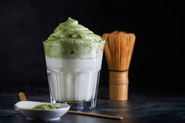 Glass of fresh dalgona coffee with matcha and Japanese tea ceremony accessories consisting of bamboo whisk called chasen and traditional scoop named chashaku placed on gray surface - ADSF33604