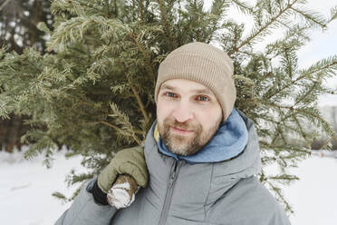 Mann mit Strickmütze trägt Tannenbaum auf der Schulter - EYAF01913