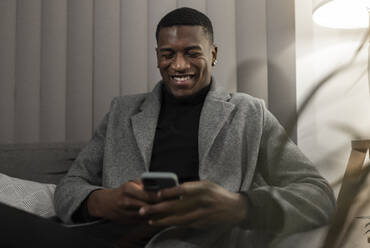 Smiling young man using smart phone sitting on sofa at home - JPTF01039