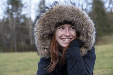 Lächelnde junge Frau mit warmem Pelzmantel mit Kapuze schaut in die Natur - LBF03618