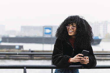 Smiling young woman with curly hair holding smart phone leaning on railing - PNAF02944