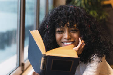 Smiling young woman reading novel at cafe - PNAF02948