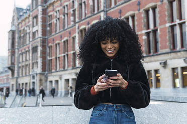 Fröhliche, modische, junge, lockige Frau mit Textnachrichten auf einem Smartphone in der Stadt - PNAF02952