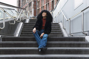 Fröhliche junge Frau mit lockigem Haar sitzt auf einer Treppe in der Stadt - PNAF02953