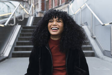 Happy young curly haired woman in front of staircase - PNAF02955