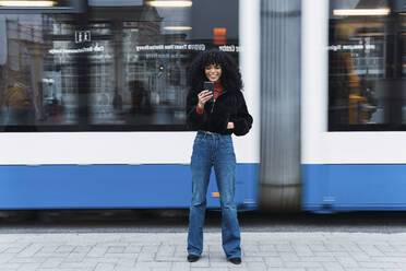 Modische junge Frau benutzt ihr Smartphone vor einer fahrenden Straßenbahn - PNAF02956